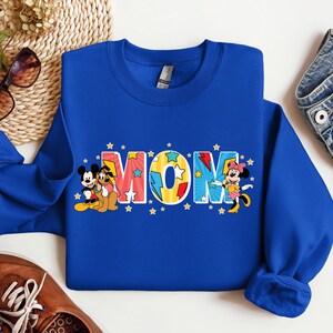 K&ouml;nnte beinhalten: Blauer Pullover mit einem bunten "MOM"-Motiv mit Mickey Mouse, Minnie Mouse und Pluto. Die Buchstaben sind gelb umrandet und haben einen Comic-Stil.