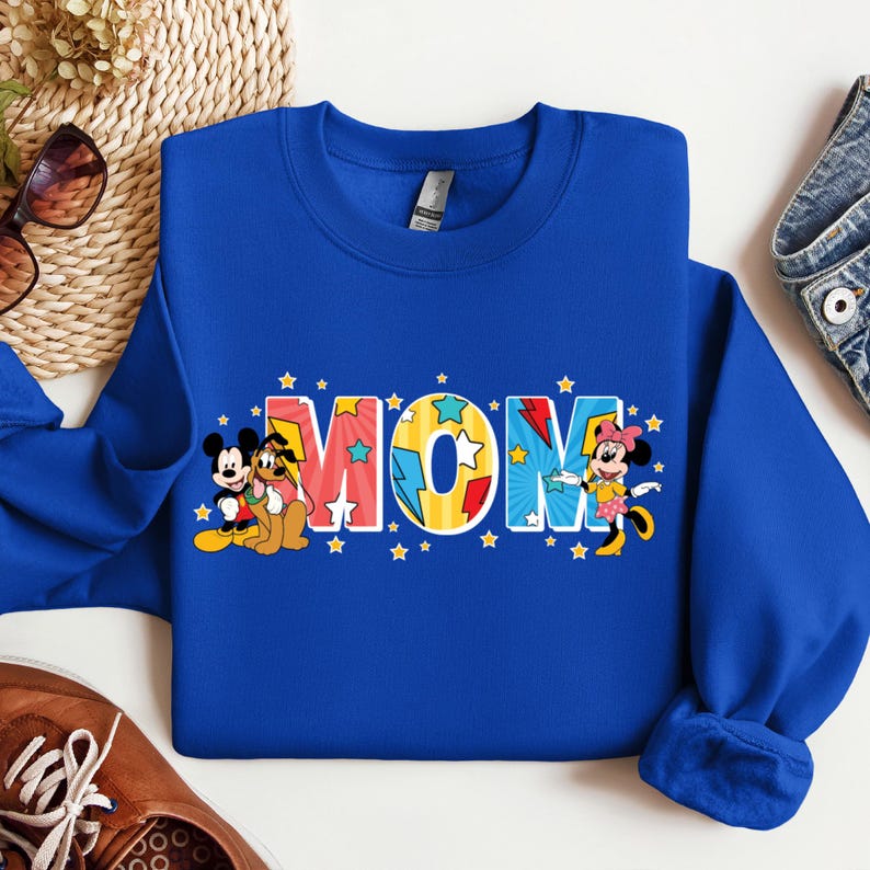 K&ouml;nnte beinhalten: Blauer Pullover mit einem bunten "MOM"-Motiv mit Mickey Mouse, Minnie Mouse und Pluto. Die Buchstaben sind gelb umrandet und haben einen Comic-Stil.