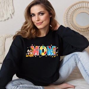 K&ouml;nnte beinhalten: Schwarzer Pullover mit einem bunten "MOM"-Motiv mit Mickey Mouse, Pluto und Minnie Mouse. Die Buchstaben sind mit gelben Sternen verziert.