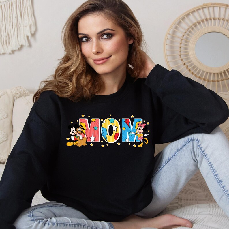K&ouml;nnte beinhalten: Schwarzer Pullover mit einem bunten "MOM"-Motiv mit Mickey Mouse, Pluto und Minnie Mouse. Die Buchstaben sind mit gelben Sternen verziert.