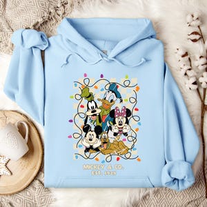 May include: Light blue hooded sweatshirt with a graphic print of Mickey Mouse, Minnie Mouse, Donald Duck, Goofy, and Pluto surrounded by colorful Christmas lights. The text "Mickey & Co. Est. 1928" is printed below the graphic.