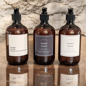 Puede incluir: Tres botellas de vidrio ámbar con tapas de bomba negras y etiquetas que dicen "All Natural Hand Soap" con los ingredientes y el tamaño indicados. Las botellas están sobre una encimera de mármol.