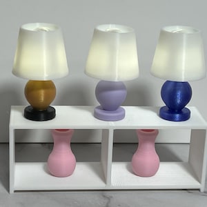 May include: Three miniature table lamps with white conical shades and spherical bases in gold, lavender, and blue, positioned on a white rectangular shelf unit. Two small pink vases are placed in the open compartments beneath the lamps.