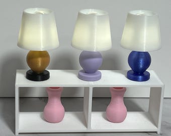 May include: Three miniature table lamps with white conical shades and spherical bases in gold, lavender, and blue, positioned on a white rectangular shelf unit. Two small pink vases are placed in the open compartments beneath the lamps.