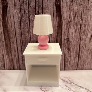 May include: A miniature white nightstand with a drawer and open shelf, topped with a small lamp. The lamp has a white shade and a pink base. The background is a wooden wall.
