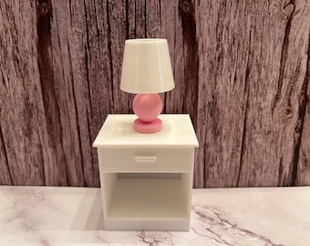 May include: A miniature white nightstand with a drawer and open shelf, topped with a small lamp. The lamp has a white shade and a pink base. The background is a wooden wall.