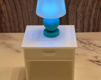 May include: A miniature teal and white lamp with a glowing blue light, resting on a small white nightstand. The lamp has a rounded base and a lampshade. The nightstand has a drawer and a rectangular shape.