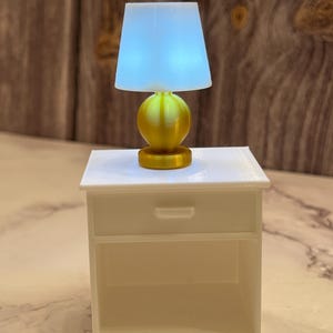 May include: A miniature gold-colored lamp with a light blue lampshade sits atop a small white nightstand. The lamp has a spherical base and a cylindrical base. The nightstand has a drawer and an open shelf.