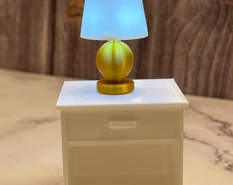 May include: A miniature gold-colored lamp with a light blue lampshade sits atop a small white nightstand. The lamp has a spherical base and a cylindrical base. The nightstand has a drawer and an open shelf.