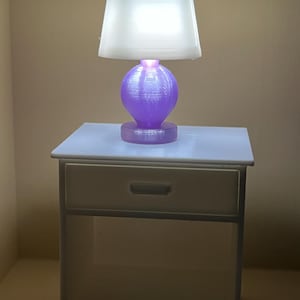 May include: A miniature white nightstand with a single drawer and an open shelf below. Sitting on top is a small table lamp featuring a translucent white tapered shade and a round, textured purple base that emits a soft, warm light.