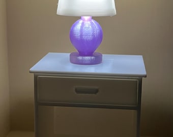 May include: A miniature white nightstand with a single drawer and an open shelf below. Sitting on top is a small table lamp featuring a translucent white tapered shade and a round, textured purple base that emits a soft, warm light.