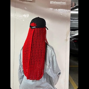 May include: A person wearing a red crocheted scarf over their head and a black baseball cap with the word "Jordan" on it. The person is wearing a light blue denim jacket.