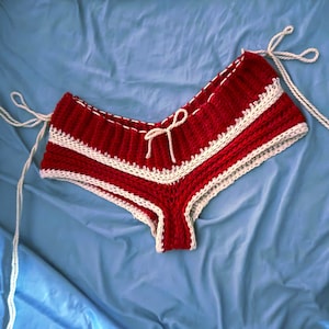 May include: Handmade red and white crochet shorts. The shorts feature a red body with white trim and accents. White ties are on each side. The shorts are made of a soft, textured yarn.