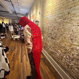 May include: A person wearing a red, open-weave scarf and hood combination. The scarf drapes down the back, and the person is wearing a patterned shirt and camouflage pants. The background features a brick wall and wooden floor.