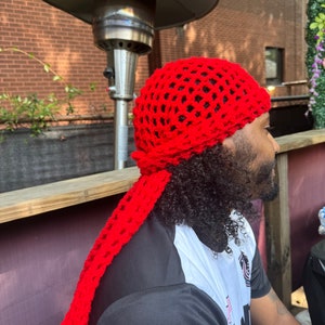 以下が含まれることがあります： 長い尻尾が付いた赤い編み物帽子。帽子は緩く開いたステッチで作られており、正方形の形をしています。尻尾はスカーフとして着用するのに十分な長さです。