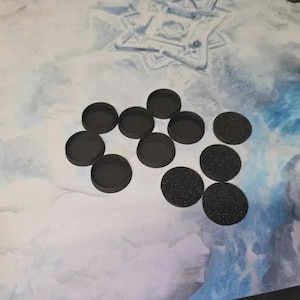 May include: A collection of black, circular bases for miniature figures. Some bases are smooth, while others have a textured, granular surface. The bases are arranged on a light blue and white patterned surface, likely a gaming mat.