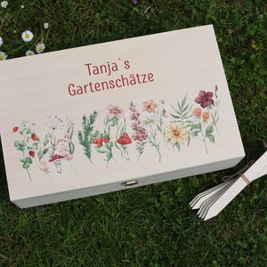 Op de afbeelding: Een lichtgekleurde houten kist met een bloemenontwerp en de tekst "Tanja's Gartenschätze". De kist is versierd met aquarel illustraties van bloemen, paddenstoelen en bladeren. Een bundel witte plantenlabels ligt op het gras.