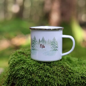 Può includere: Tazza di smalto bianco con una scena di foresta e il nome "Nils" stampato su di essa. La tazza è appoggiata su un letto di muschio verde.