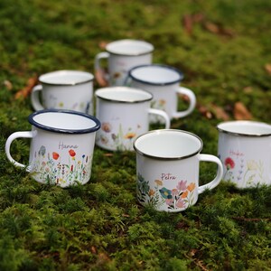 May include: Eight white enamel mugs with blue rims and floral designs. Each mug has a different name written on it, including Hanna and Petra.