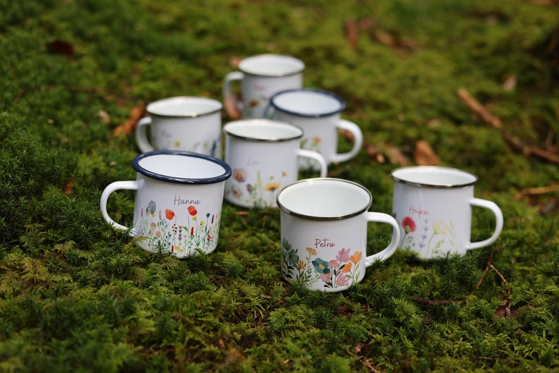 May include: Eight white enamel mugs with blue rims and floral designs. Each mug has a different name written on it, including Hanna and Petra.