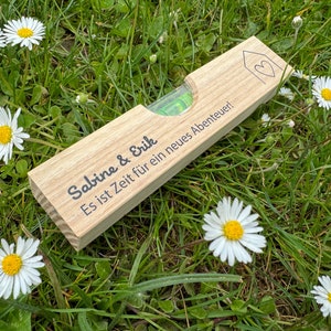 Op de afbeelding: Een houten waterpas met een groene bel en een hartvormig huisontwerp. De waterpas is gegraveerd met de tekst "Sabine & Erik Es ist Zeit für ein neues Abenteuer!"