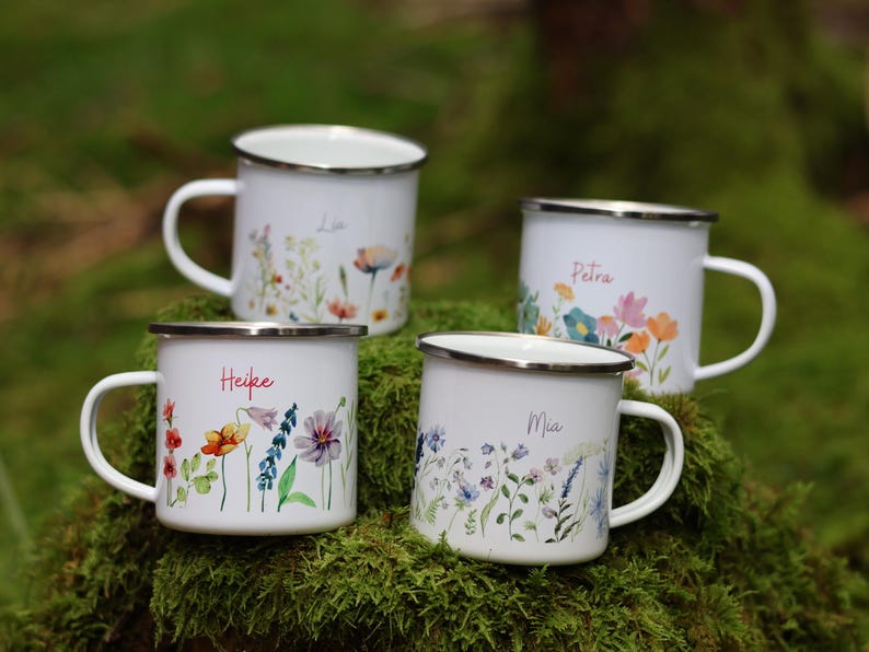 May include: Four white enamel mugs with floral designs and names "Lia", "Heike", "Petra", and "Mia" printed on them. The mugs are sitting on a bed of green moss.