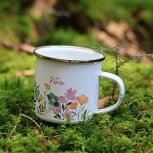 May include: White enamel mug with a floral design and the name "Petra" printed on the front. The mug is sitting on a bed of green moss.