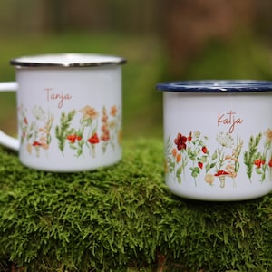 May include: Two white enamel mugs with floral designs and names "Tanja" and "Katja" printed on them. The mugs are sitting on a bed of green moss.