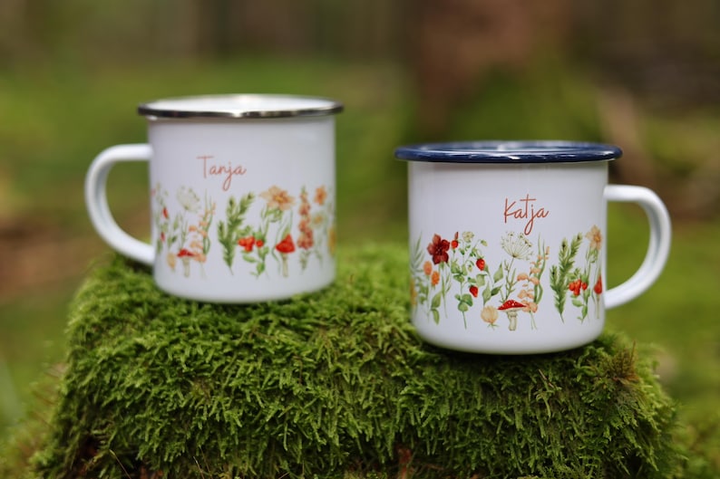 May include: Two white enamel mugs with floral designs and names "Tanja" and "Katja" printed on them. The mugs are sitting on a bed of green moss.
