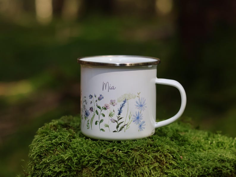May include: White enamel mug with a floral design and the name "Mia" printed on the front. The mug is sitting on a bed of green moss.