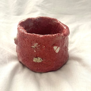 May include: A small, red, textured ceramic planter with white flecks. The planter has a rounded, irregular shape.