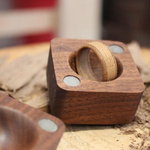 May include: A wooden ring with a light brown band rests in a square, dark brown wooden holder. The holder has two small, round, light-colored accents. Another similar holder is in the foreground, with wood shavings scattered around.
