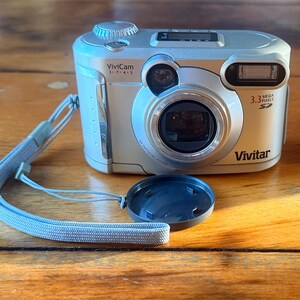 May include: Silver Vivitar digital camera with a lens cap and a light blue wrist strap. The camera has a 3.3-megapixel resolution and a 3x optical zoom. The camera is on a wooden surface.