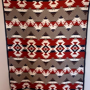 May include: A large, rectangular blanket with a geometric pattern in red, white, gray, and navy blue. The design features repeating diamond and triangle shapes. The blanket is hanging on a wall.