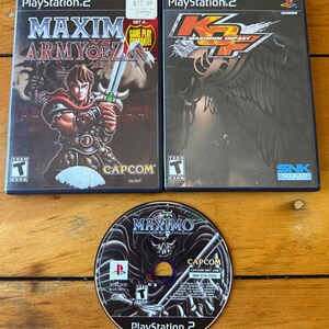 May include: Two PlayStation 2 game cases and one game disc. The left case is Maximo vs. Army of Zin, the right case is KOF Maximum Impact, and the disc is for Maximo vs. Army of Zin. Each case features a Teen ESRB rating and game title text.