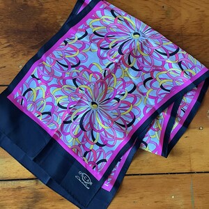 May include: A pink and black silk scarf with a white background and a floral pattern. The scarf has a navy blue border and the text "OSCAR de la Renta" in white.
