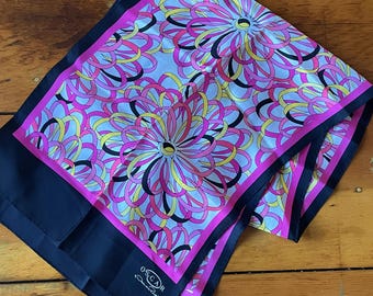 May include: A pink and black silk scarf with a white background and a floral pattern. The scarf has a navy blue border and the text "OSCAR de la Renta" in white.