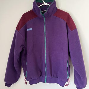 May include: A purple fleece jacket with a high collar and a full-length teal zipper. The jacket has burgundy shoulder panels and teal accents on the pockets. A small blue and white logo is on the chest.