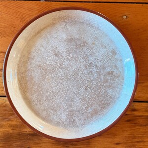 May include: A round ceramic plate with a cream-colored center and a brown rim. The center has a crackle pattern, and the rim is a darker brown. The plate is likely for serving food.