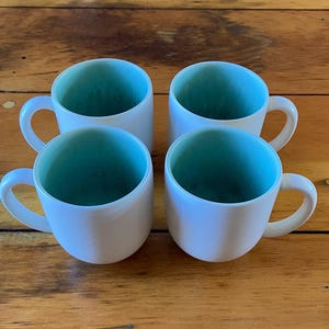 May include: Four white ceramic mugs with a teal blue interior. The mugs have a rounded shape and a handle on the side.