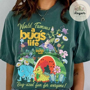 May include: A green t-shirt with a vintage-style graphic of characters from the movie 'A Bug's Life'. The graphic features the text 'World Famous A Bug's Life' and includes images of various bugs, including a ladybug, a caterpillar, and an ant. The graphic also includes the text 'Bug-sized fun for everyone!'