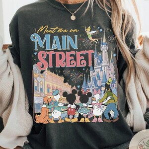 May include: Dark gray sweatshirt with a colorful graphic featuring Disney characters, a castle, fireworks, and the text "Meet me on Main Street." The design includes Mickey and Minnie Mouse, Donald Duck, and Goofy.