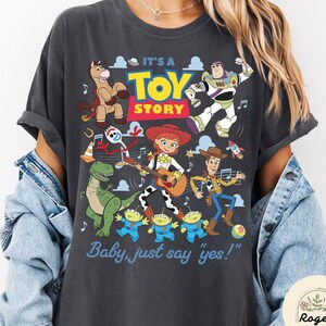 May include: A charcoal gray short-sleeve t-shirt featuring a colorful graphic print of Toy Story characters including Woody, Buzz Lightyear, Jessie, Rex, Bullseye, Forky, and the Aliens, with the text IT'S A TOY STORY and Baby, just say yes!