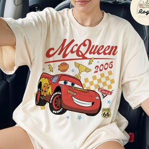 May include: Cream-colored t-shirt featuring a graphic of Lightning McQueen from the movie Cars. The design includes the text "McQueen 2006" in red, a checkered flag, and Route 66 signage. The shirt has a relaxed fit.