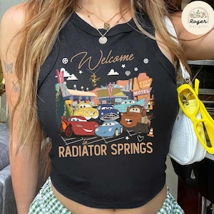 May include: Black tank top featuring a colorful graphic of characters from the movie Cars, with the text "Welcome to Radiator Springs." The design includes various cars and the Cozy Cone Motel.