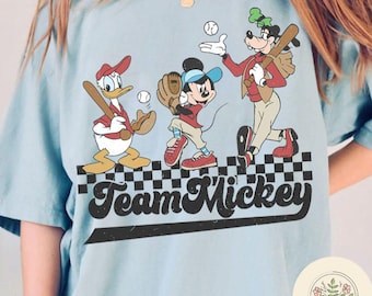 May include: A light blue t-shirt with a graphic of Mickey Mouse, Donald Duck, and Goofy playing baseball. The text "Team Mickey" is printed in black on a checkered background.