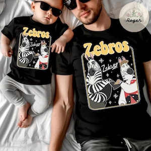 Pu&ograve; includere: T-shirt nere con la scritta "Zebros" in giallo, con un disegno di zebra dei cartoni animati. Una maglietta &egrave; indossata da un bambino, l'altra da un adulto. Entrambe le magliette hanno lo stesso disegno.