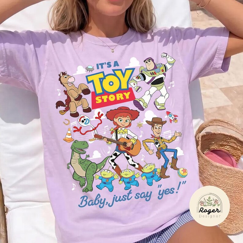 May include: A light purple t-shirt featuring a colorful graphic print of Toy Story characters including Buzz Lightyear, Woody, Jessie, Bullseye, Rex, Forky, and three aliens. The text reads IT'S A TOY STORY and Baby, just say "yes!" with musical notes.