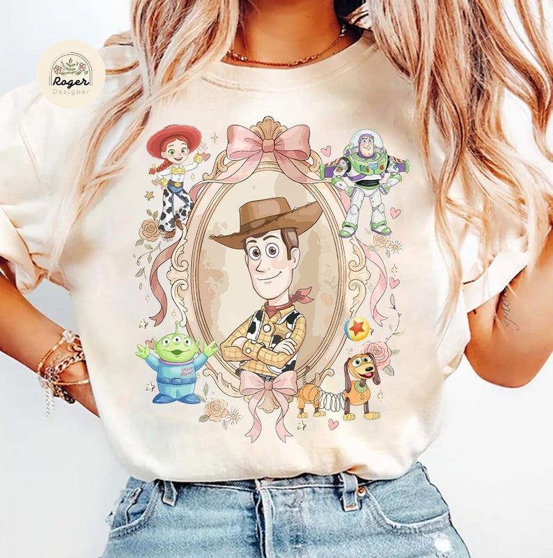 May include: Cream-coloured t-shirt featuring a framed illustration of Woody from Toy Story, surrounded by other characters like Jessie, Buzz Lightyear, and the Little Green Men. The design includes pink ribbons, roses, and a slinky dog.