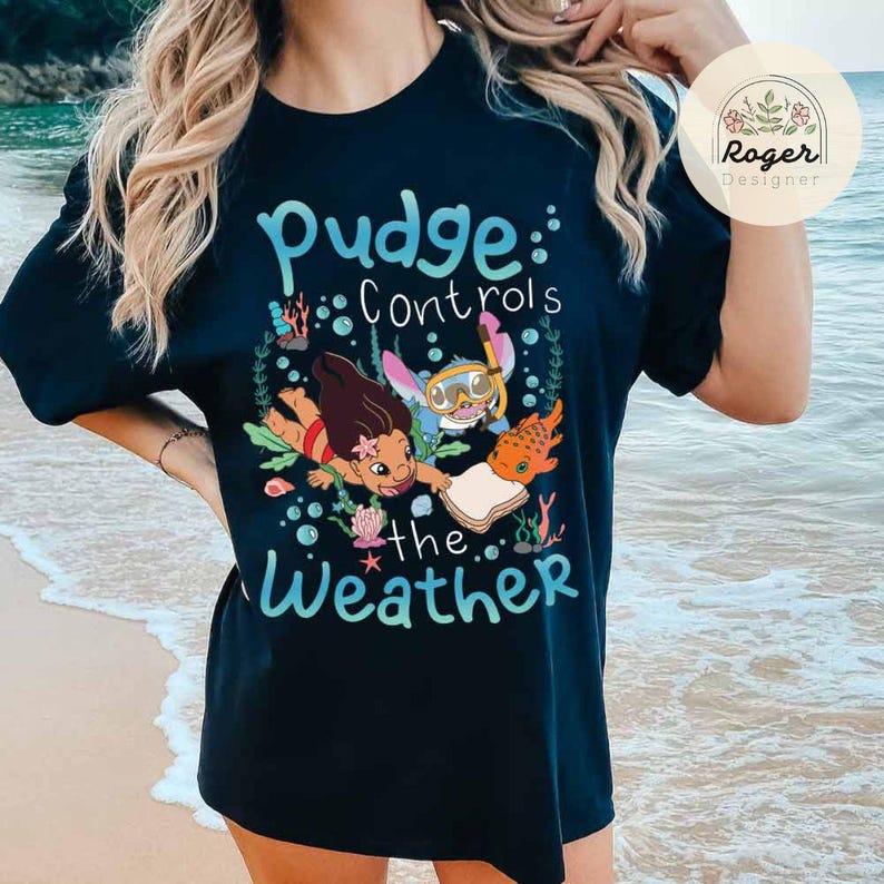 May include: Black t-shirt featuring a colorful graphic with the text "Pudge Controls the Weather." The design includes cartoon characters and aquatic elements. The shirt is a crew neck style.